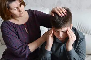 A woman is placing her hand on a boy's head while sitting on a couch, possibly trying to comfort him.