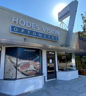 A front view of a building with a sign that reads 'Hodes Vision Optometry', featuring a large window displaying sunglasses and a logo.