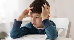 Image of a young boy looking stressed while sitting at a desk and holding his head with his hands.