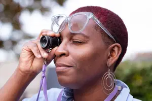 Person with short hair using a small magnifying device while wearing oversized glasses, outdoors.
