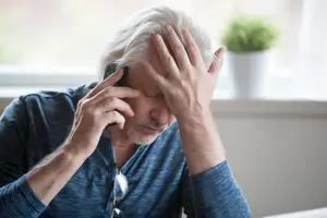 Man with gray hair holding phone, hand on forehead, looking stressed, indoors.