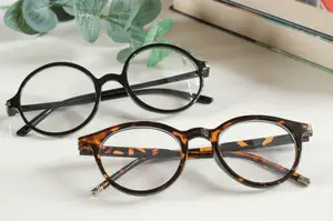 Two pairs of glasses with round lenses and black frames, one with tortoiseshell patterns, placed on a white surface next to a book and green leaves.