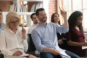 A group of people in a classroom with a man raising his hand and a woman in glasses holding a pen.