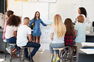 High school students sit at a table and listen to a teacher in a classroom.