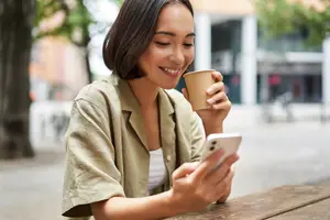 A woman smiling while looking at her phone and holding a coffee cup.