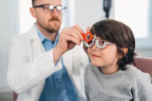 An optometrist adjusts a young child's glasses while they sit in a clinic.