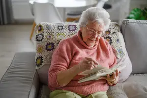 An elderly woman wearing glasses and a pink sweater is sitting on a couch and reading a book.