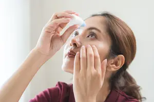 A woman is putting eye drops in her eye while looking in a mirror.