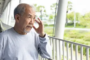 An elderly man is standing on a balcony with his hand on his ear, looking out towards a grassy area with trees and a pathway in the background.