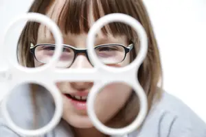 A young girl wearing glasses is looking through a white circular object with multiple holes, possibly a visual aid.