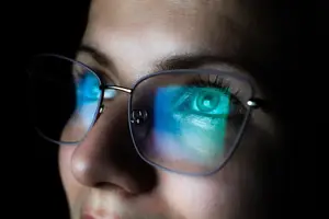 A woman with glasses reflecting light, focusing on her left eye.