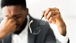 A close-up of a man holding glasses in his hand and covering his eyes with his other hand, probably in a meeting.