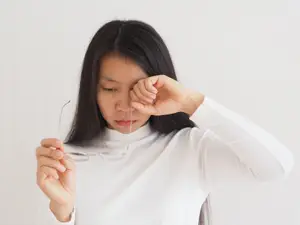 A woman wearing a white long-sleeve shirt is holding a pair of glasses and touching her eye with her finger.
