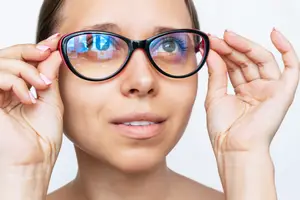 A woman wearing glasses with a reflection of her eyes, holding the glasses with both hands and looking upward.