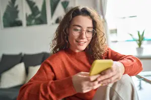 A woman wearing glasses and a red sweater is smiling and looking at her phone.