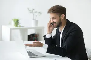 A man in a suit is sitting at a desk with a laptop, talking on a phone.
