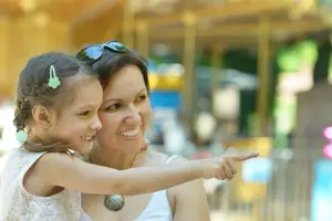 A smiling woman and a young girl point at something in an amusement park, both wearing accessories.