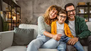 A family of three, including a woman, a man, and a child, is sitting on a couch and smiling for a photo.