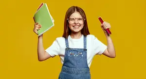 A young girl with glasses and denim overalls is holding a book and a pencil, smiling and posing for a photo.
