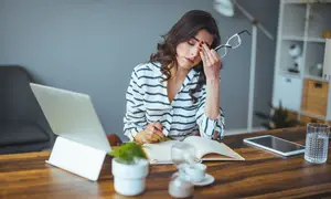 Woman with a headache sitting at a desk with a laptop and book