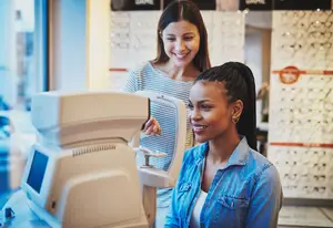 A woman is checking her eye vision using an eye examination machine at an optical shop with another woman smiling in the background.