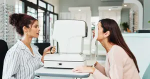 Two women are in an eye care clinic, one looking at an eye chart while the other is examining her eyes with a device.