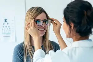 A woman is trying on glasses at the optometrist with a smiling doctor assisting her