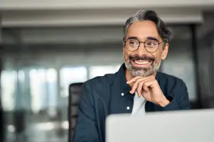 Smiling middle-aged man with gray hair and beard wearing glasses and a blue shirt sitting at a desk with a laptop in front of him.