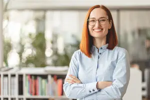 A woman in a blue shirt and glasses is smiling and standing in an office room with bookshelves and plants in the background.