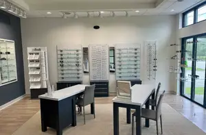 A modern optical store with white countertops, black tables, and a glass display case with glasses. There are also chairs and mirrors.