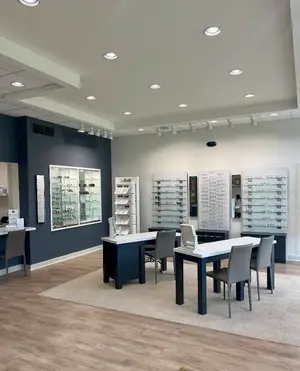An optical shop with blue walls, white ceiling, wooden floor, and several tables and chairs. The shop has a modern design with various eyeglass frames on display, and a person is standing behind the counter.