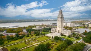 An aerial view of the Louisiana State Capitol building in Baton Rouge, Louisiana, surrounded by a lush green lawn and trees, with a river and cityscape in the background.