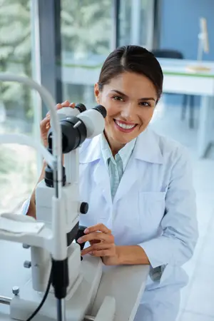 An eye doctor wearing a white lab coat smiles while using an ophthalmoscope on a patient