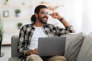 A man sitting on a couch with a laptop on his lap, smiling and adjusting his glasses