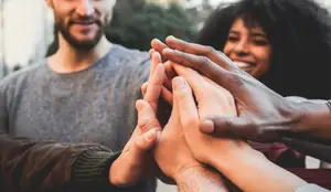 A group of people standing together with their hands stacked on top of each other, possibly for a team-building activity or meeting, while smiling and looking at the camera.