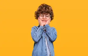 A young boy with glasses covering his mouth with his hands while smiling in front of a yellow background