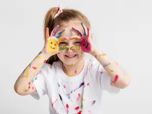 A young girl with her hands covered in multicolored paint stands against a white background, smiling and posing for a photo.