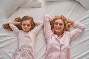 A young girl and her mother are laying on a bed, both are wearing pink striped pajamas and pink sunglasses.