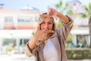 Portrait of a smiling woman with glasses standing outdoors, gesturing with her hands near a building and palm trees in the background.