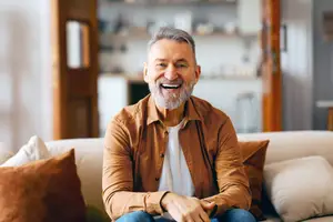 A smiling man in a brown jacket sits on a couch with his hands clasped together, next to a shelf and door.