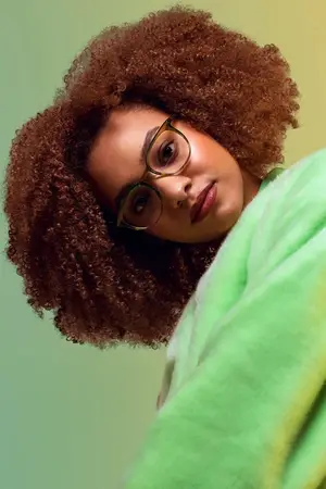 A woman with curly hair and green glasses poses for a photo wearing a green fur coat.