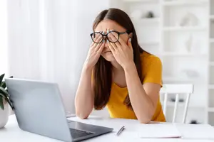 A woman in a yellow shirt wearing glasses is sitting in front of a desk with a laptop, a book, and a pen, with her hands covering her eyes.