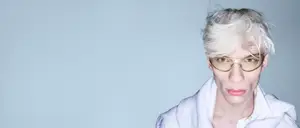 A person with white hair wearing glasses and a white shirt against a blue background