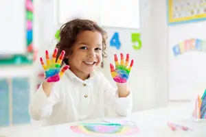 A child with rainbow colored hands sitting at a table smiling at the camera