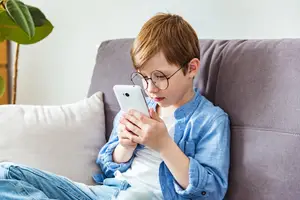 A young boy wearing glasses and sitting on a couch while holding a phone