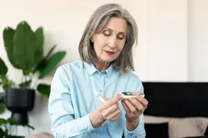 An elderly woman with gray hair is sitting on a couch and holding a glucometer in her hands, checking her blood sugar level. She is wearing a blue shirt and seems to be in a room with a couch, a black headboard, and some potted plants.