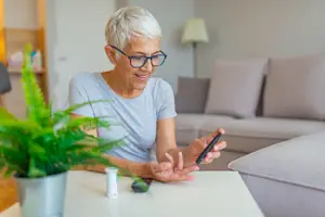 An older woman with short gray hair and glasses sits on a couch and checks her blood sugar with a glucometer.