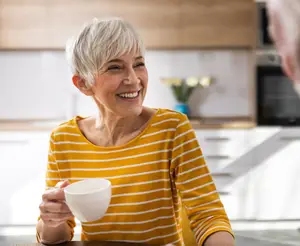 A smiling woman in a yellow striped shirt holding a cup is sitting in front of a man in a blue shirt in a kitchen.