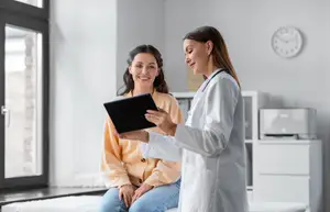 A doctor in a white coat is showing a tablet to a woman sitting on a bed in a room with white walls, a glass window, and a clock on the wall, and a cabinet with a printer and a shelf with a printer on it