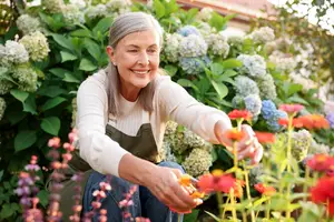 An older woman in a garden is pruning flowers with a smile on her face.